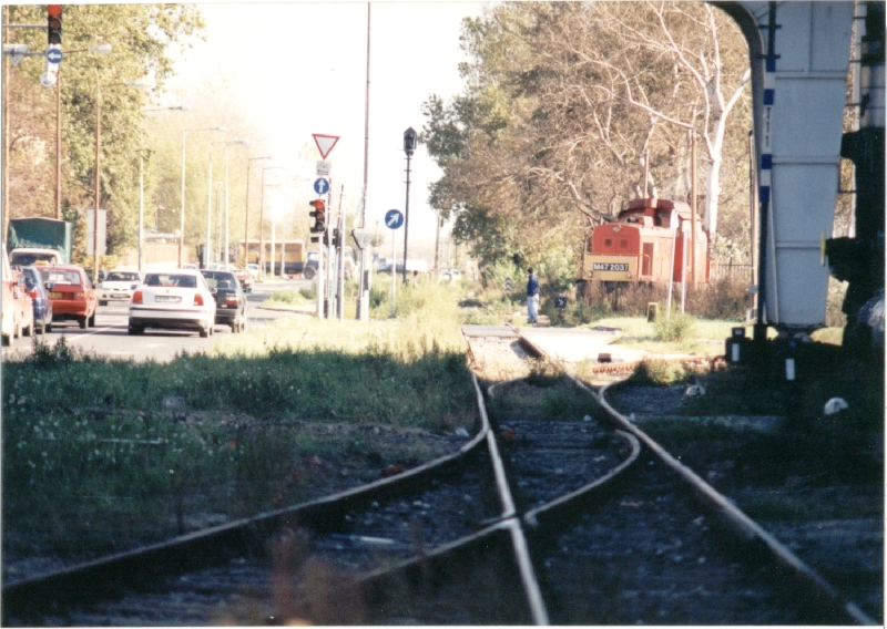 és tényleg! Pont ugyanezen a kapun halad ki egy Dacia 1998-ban. A Szerémi út ekkor már megvan, ezt 1995-ben építették ki az újbudai tehermentesítő út részeként, az iparvágányokat részben keresztezve, részben felszámolva. A képen látható gépmenet érdekessége, hogy az iparvágány-hálózatot 1997. december 31-én hivatalosan üzemen kívül helyezték, de az Épületelemgyár ezután is ragaszkodott a vasúti kiszolgáláshoz, így ritkán továbbra is jártak vonatok ebbe az irányba, mígnem 2000 körül a MÁV véglegesen felmondta a szállítási szerződést. Ma már csak a gyárak belső területein találkozhatunk sínekkel, így például a táborainkból újbudai falmászóhely udvarán. Az alsó képen a volt Keksz- és Ostyagyár területén lévő fordítókorongot láthatjuk 2006-os állapotában. (Kép és információk: http://villamosok.hu/balazs/bpvasut/ipvg/andor/index.html)